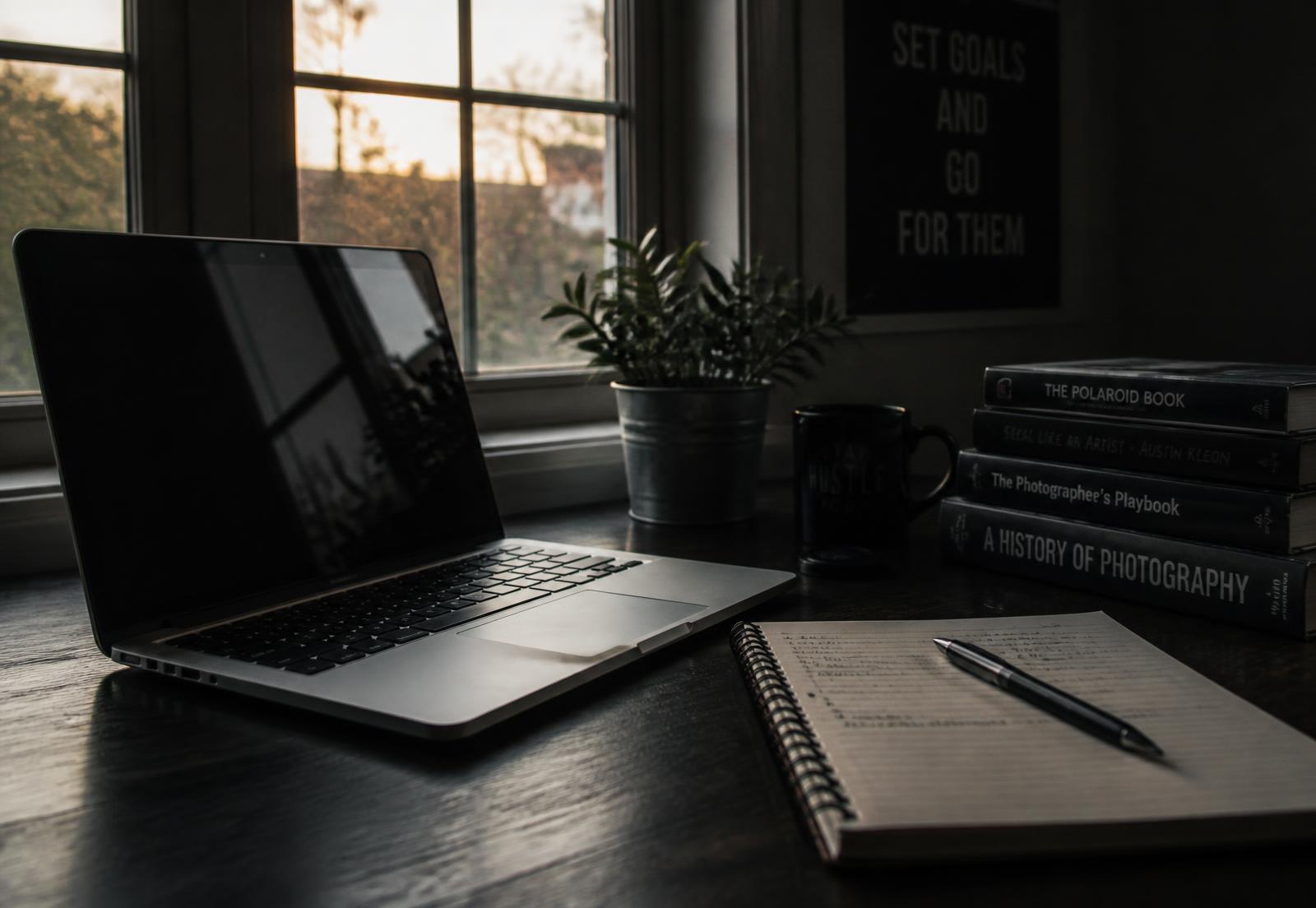Laptop, notebook and pen on a dark desk beside a bright window